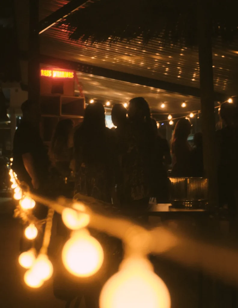 Atmospheric night photo of a group of people socializing under hanging light bulbs, with the "BASS WARRIORS" sign visible in the distance.