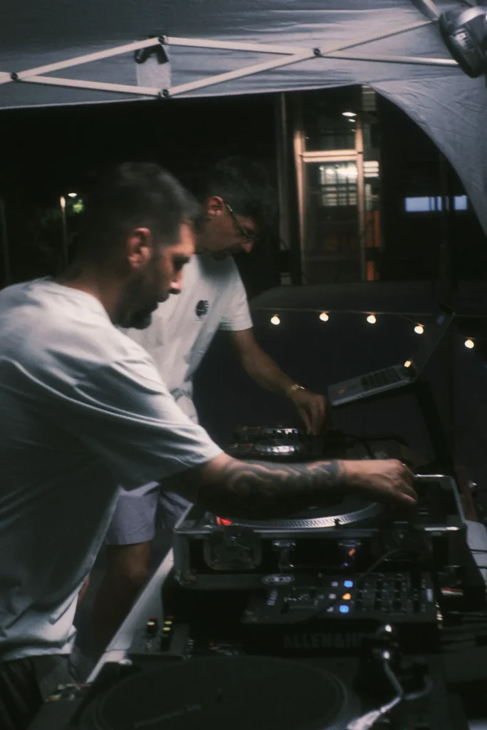 Two DJs (one with tattoos) adjusting the music on turntables and a mixer under a brightly lit tent at a night event.