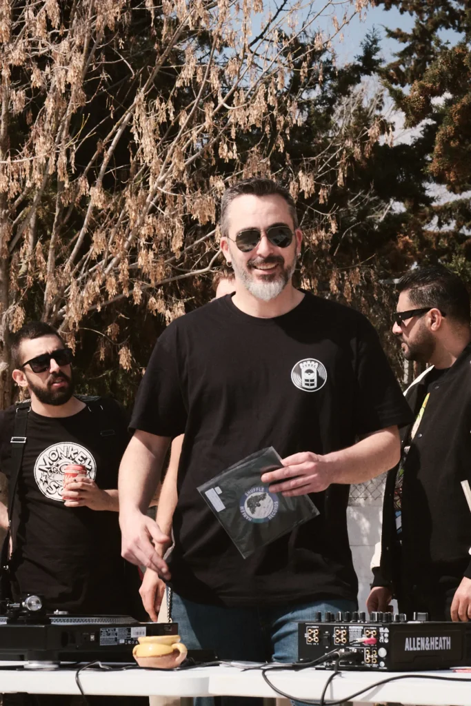 Portrait of DJ smiling, holding a vinyl record, standing next to an Allen & Heath mixer console at the Bass Warriors event.
