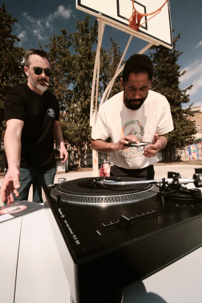 Two men interacting with a Pioneer turntable and vinyl records at an outdoor music event, with a basketball hoop in the background.