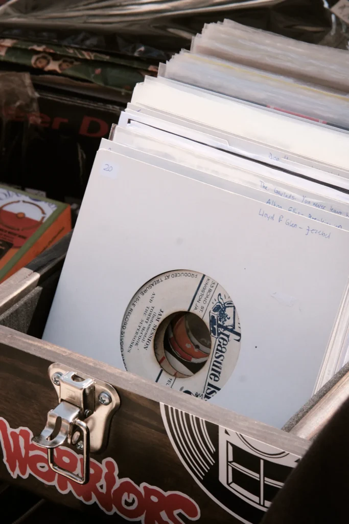 Close-up of a wooden DJ crate filled with white-sleeved 7-inch vinyl records, showing a Treasure Isle label and handwritten notes.
