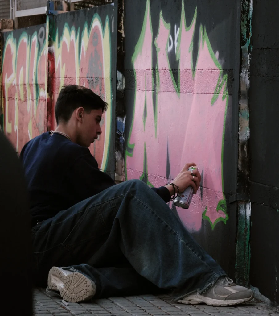 Young male graffiti artist sitting down, focused on spray painting a large pink tag on a black wall outdoors.