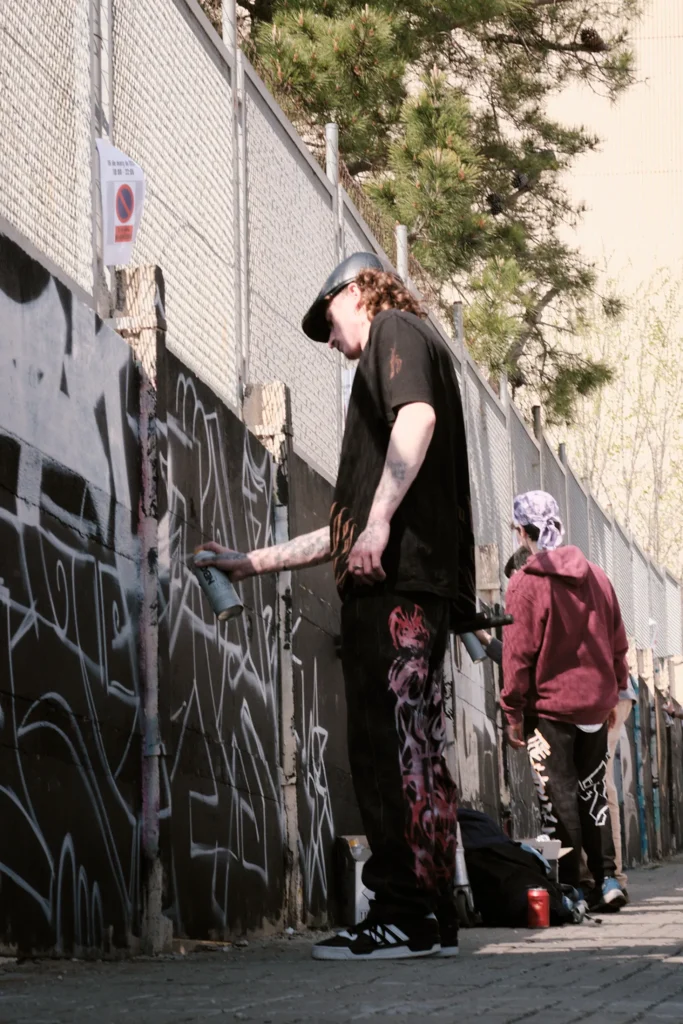 Two street artists painting black and white graffiti tags on a high urban wall next to a wire fence under sunlight.