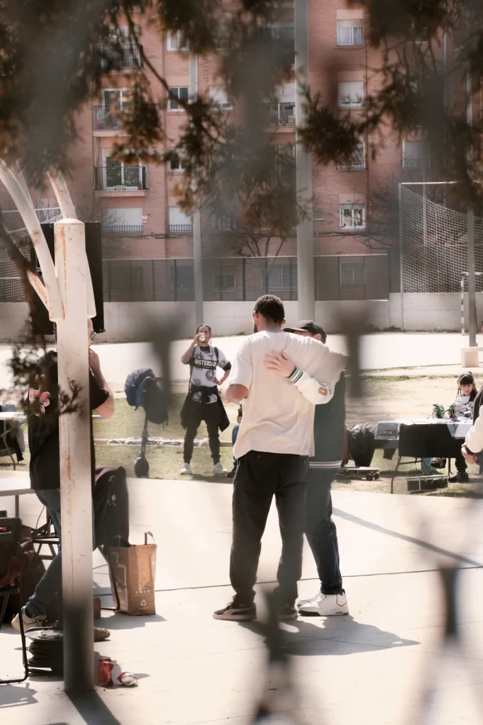 Candid atmosphere shot of people gathered outdoors at a music event, with two people embracing in the foreground.