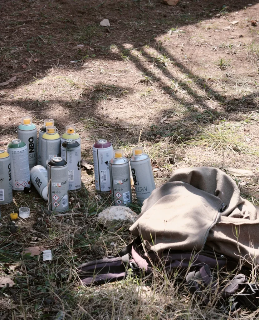 Still life of multiple colored spray paint cans (rattles) lying on the ground next to a brown jacket, detailing artist supplies.