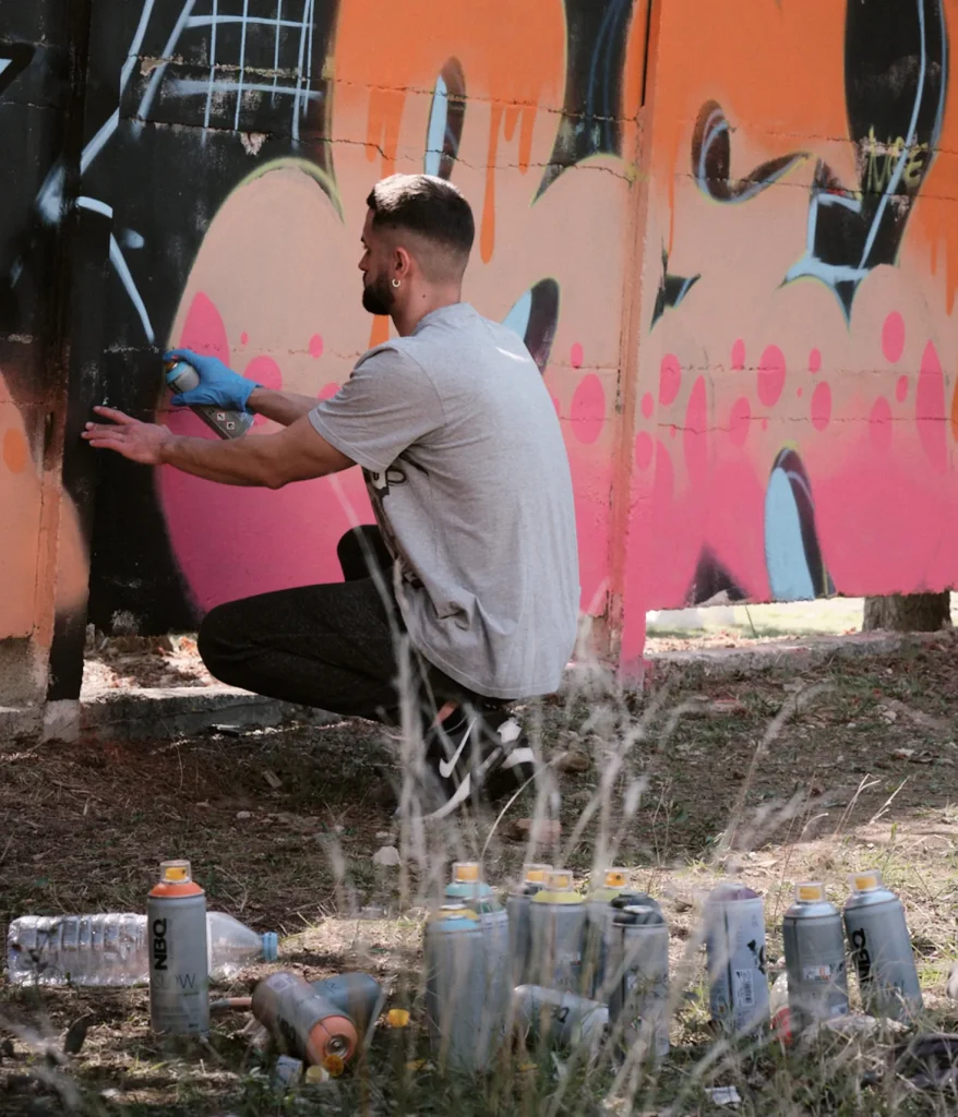 Graffiti artist squatting, painting a large-scale mural with bright pink and orange spray paint during the BassWarriors event.