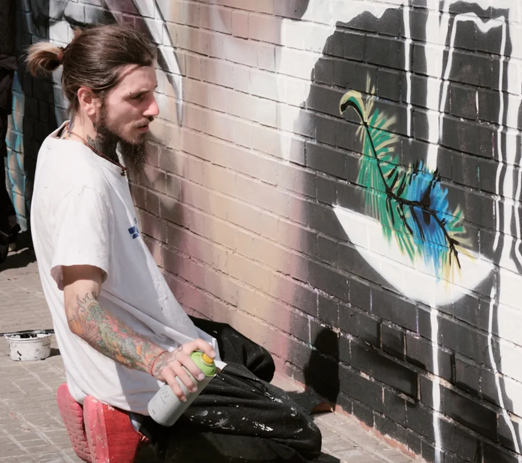 Male graffiti artist sitting against a wall, adding details with a spray can next to a large mural of an eye and a feather.