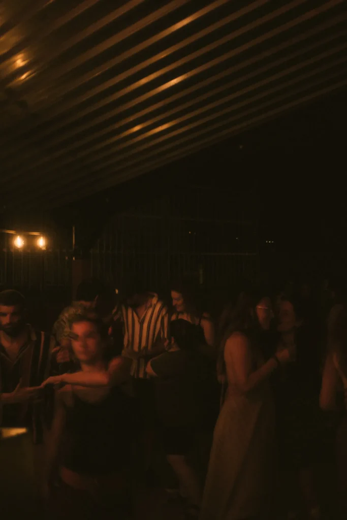 Low-light photo of a crowd of people dancing and enjoying the music under warm string lighting at night.