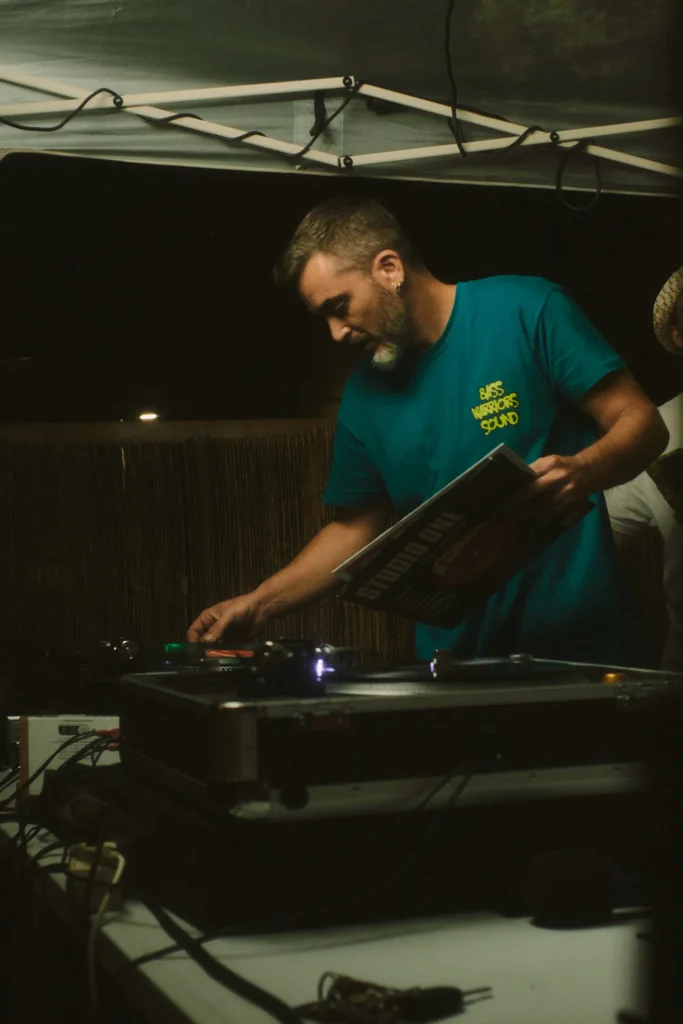 DJ in a teal shirt holding a vinyl record sleeve while selecting the next track from the turntable setup.