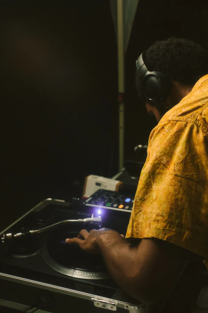 Close-up back view of a DJ wearing headphones and a yellow shirt, using a turntable during a live reggae session.