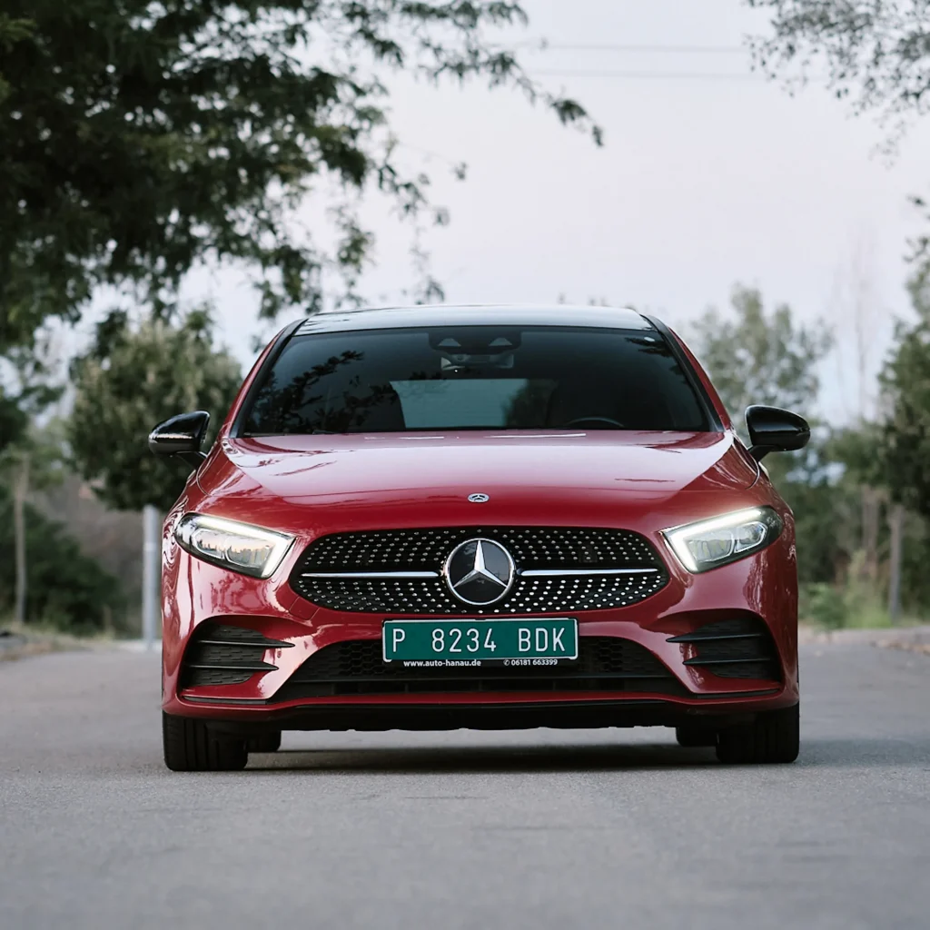 Commercial head-on view of red Mercedes A-Class car with daytime running lights on, centered on road between trees.
