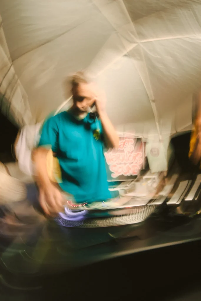 Dynamic motion-blurred image of a DJ wearing a teal Bass Warriors shirt and headphones while mixing vinyl.