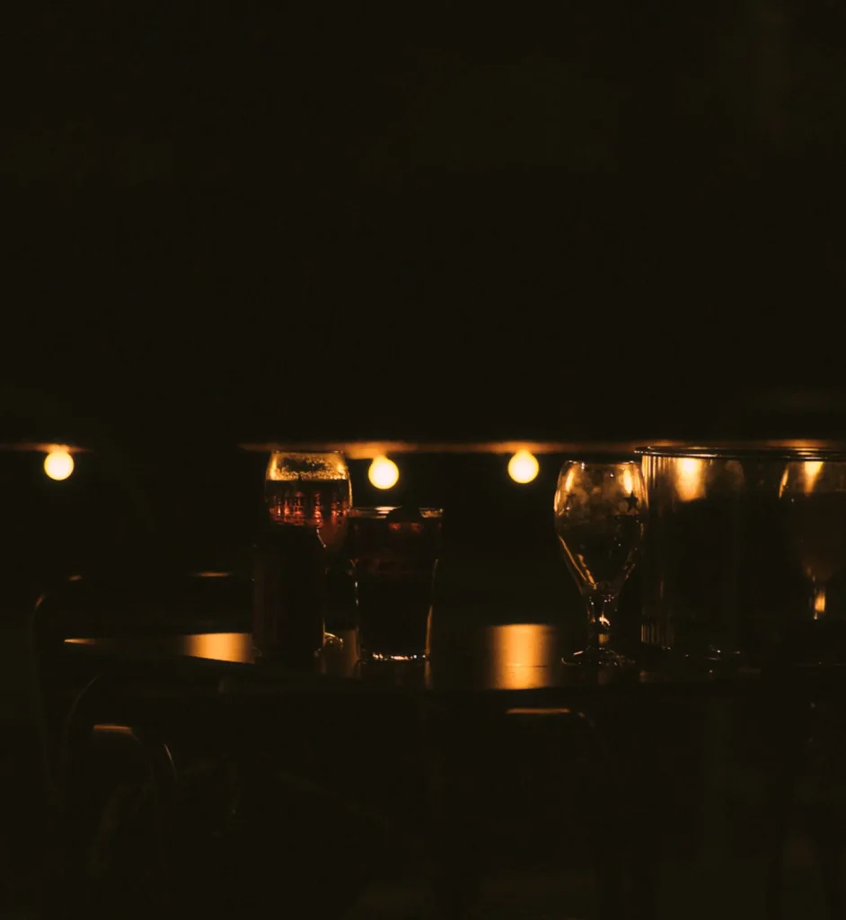 Dark, atmospheric shot of assorted glasses and drinks on a bar or table, illuminated by a string of warm fairy lights.
