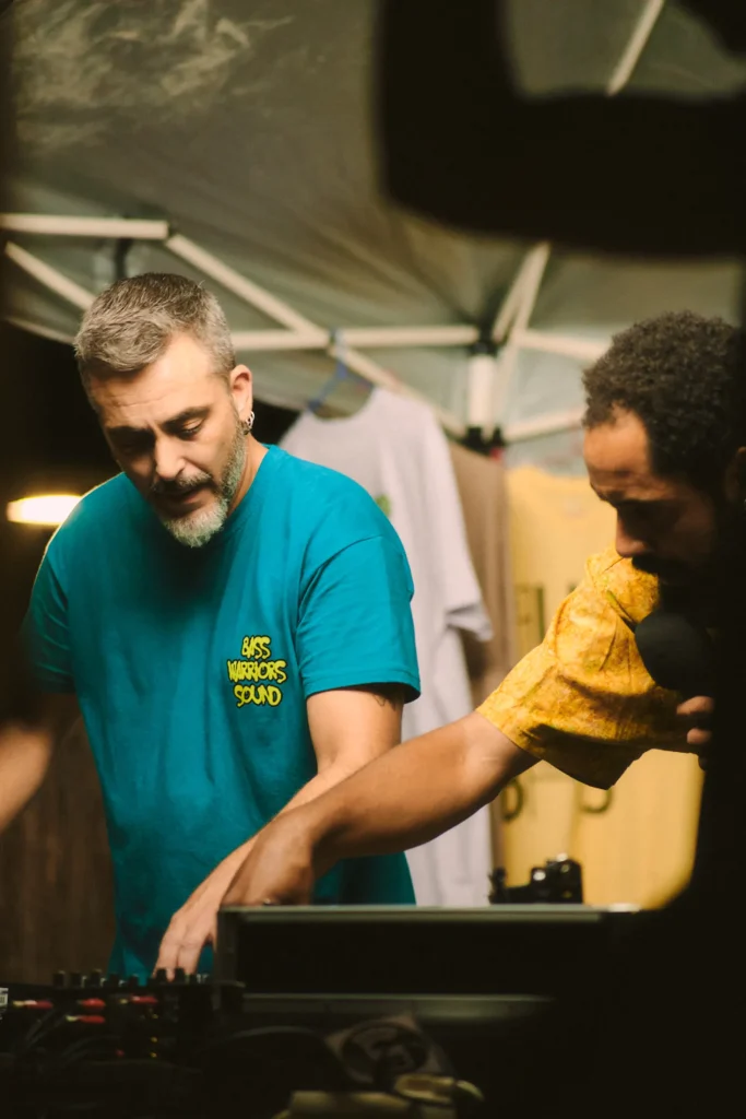 Two male DJs (one in a blue shirt, one in yellow) mixing music and interacting with the crowd at the outdoor sound system event.