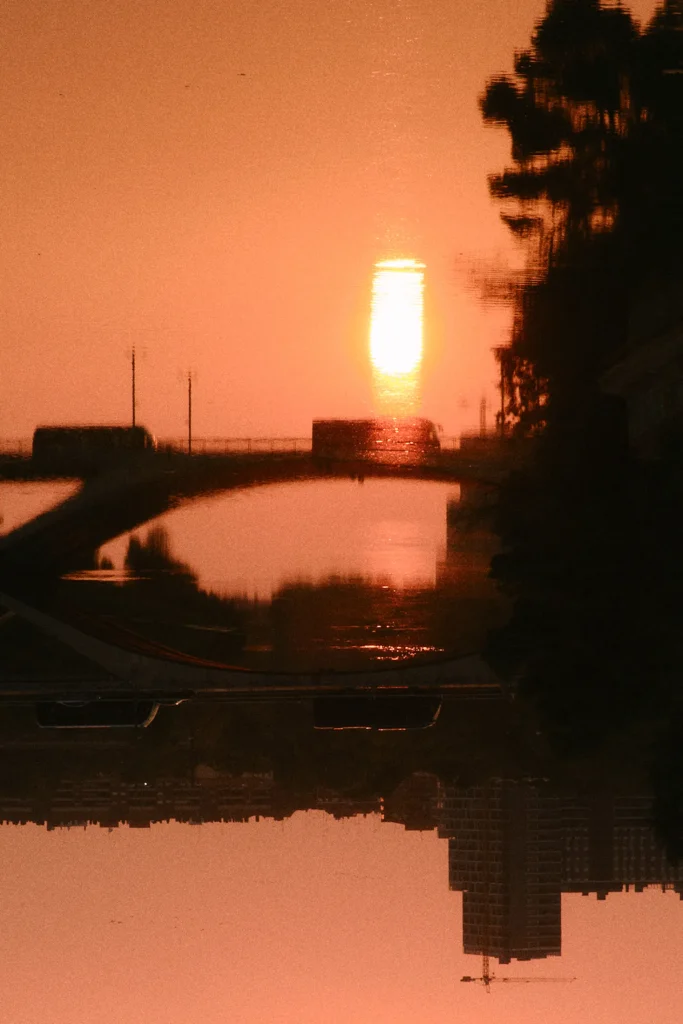 Abstract, moody urban travel photography of a city reflection on water at sunset, featuring traffic silhouettes and a glowing sun flare, creating a cinematic travel look.