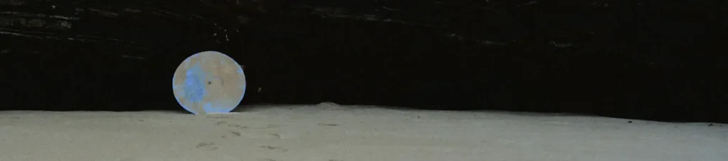 Abstract conceptual photography featuring a small, glowing moon-like disc resting on sand against a completely dark background, emphasizing a minimalist and mysterious travel atmosphere