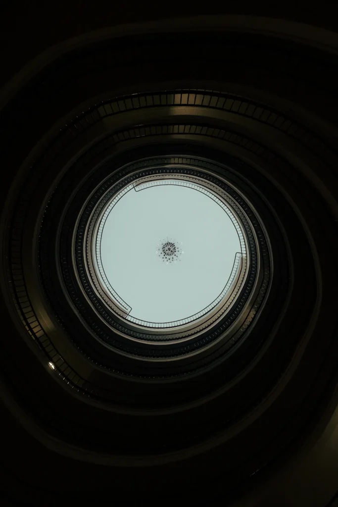 Abstract conceptual architecture photography looking up a geometric spiral staircase, emphasizing dark, circular design and minimalist patterns for high-end editorial content.