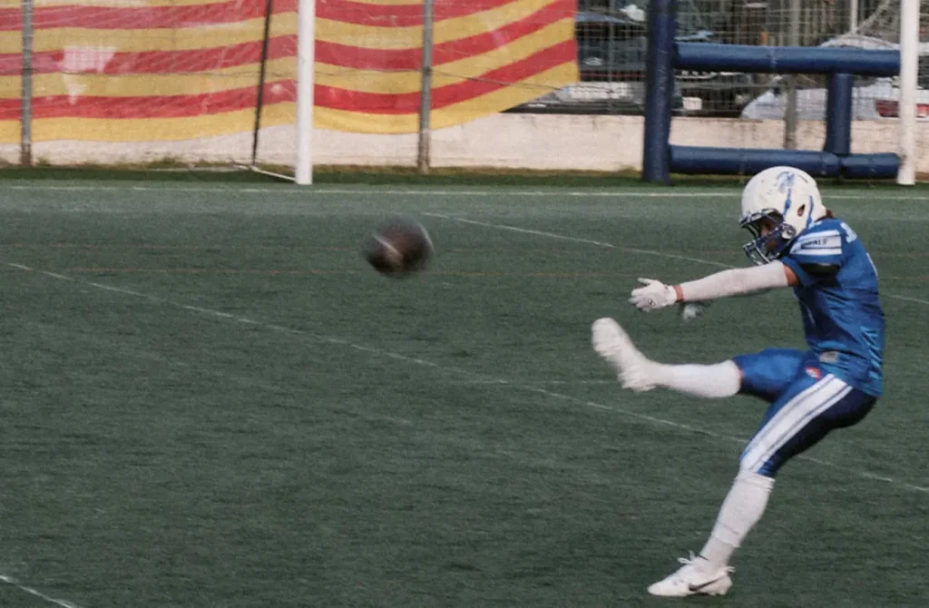 Close-up dynamic photograph of an American football player kicking a field goal, with the ball in motion.