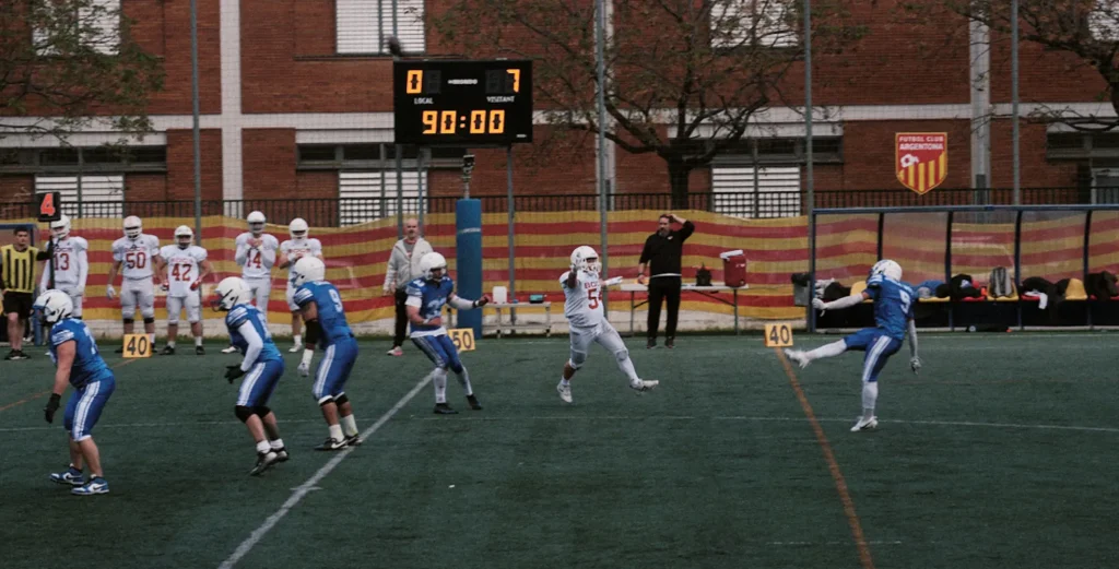 Wide action shot of an American football player kicking the ball, showing the scoreboard and opponent team lineup.