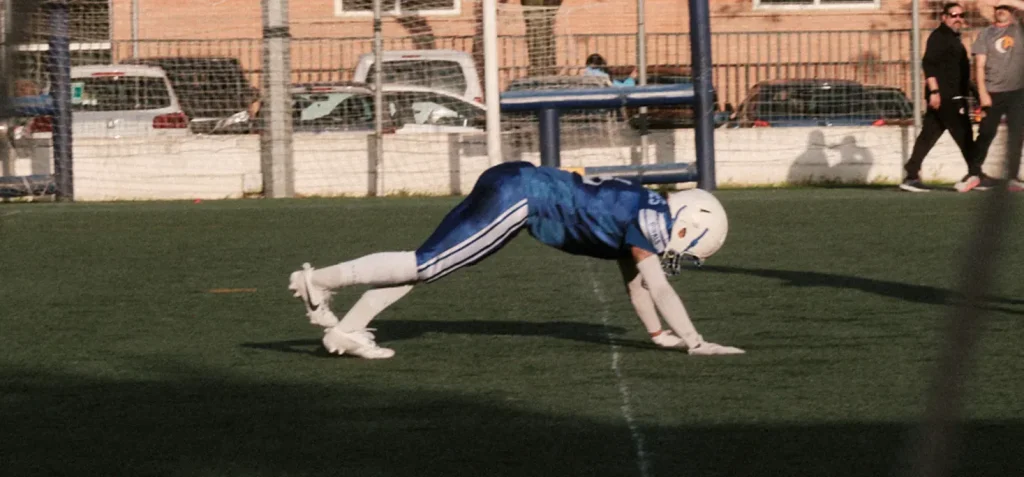 Focused image of an American football player in a blue uniform performing a low, recovery or drill posture on the field.