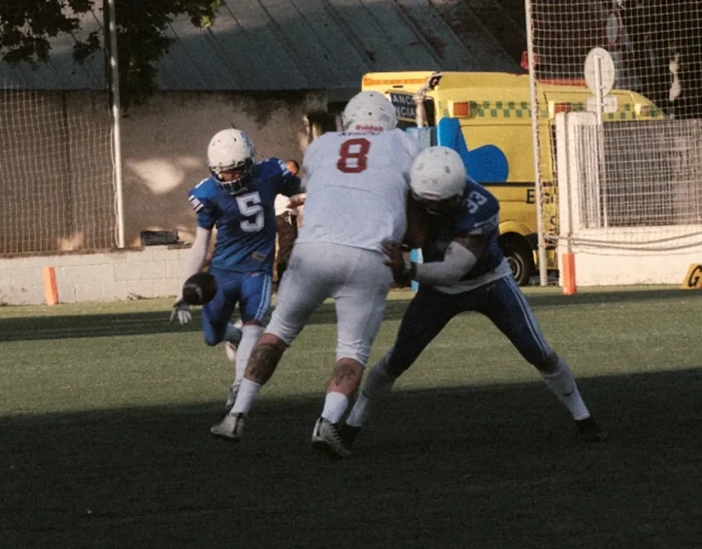 High-impact action photo of two players tackling the ball carrier, showing game intensity and safety personnel in the background.