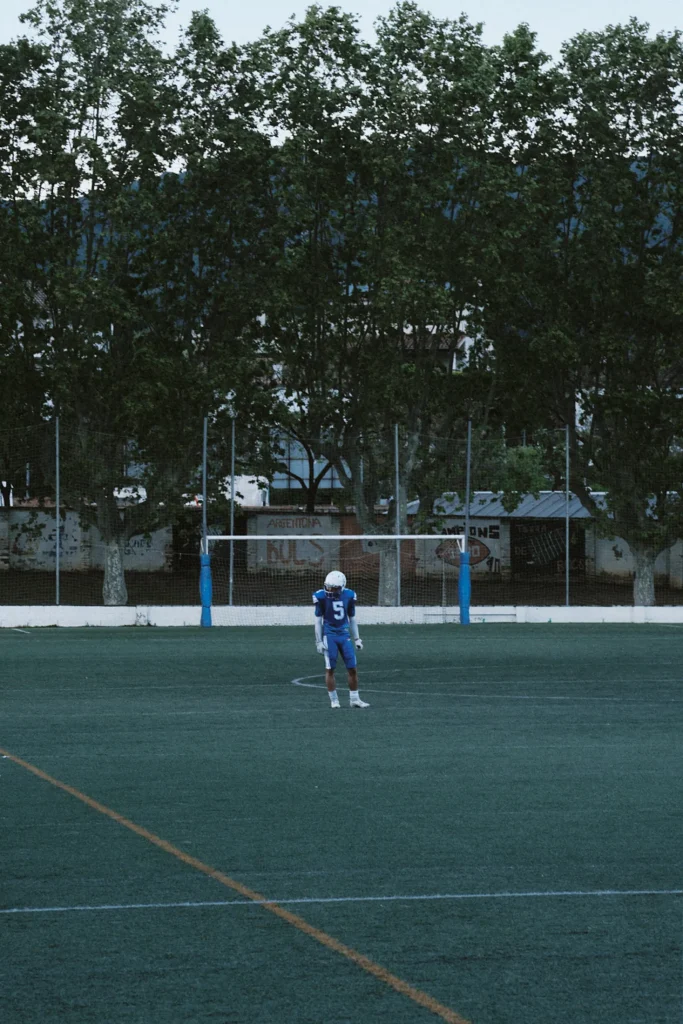 Wide, atmospheric photo of a single American football player standing alone on the field with goalposts in the background at twilight.