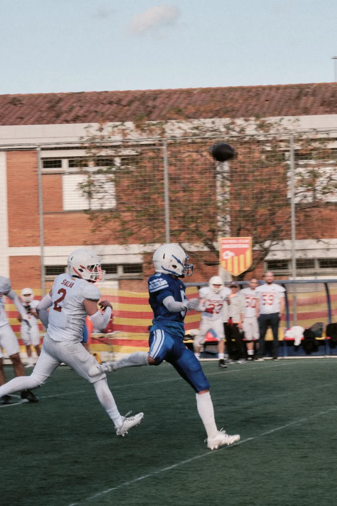 Action photo of a player kicking or punting the football, capturing the ball mid-air against the evening sky and red brick background.