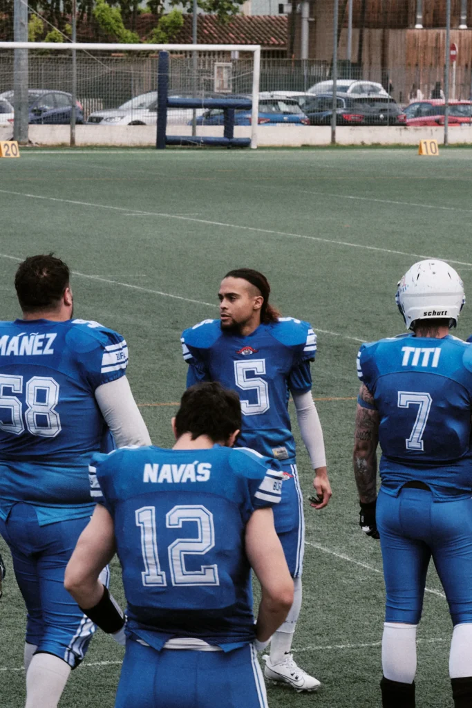 Portrait-style photo of four football players in blue jerseys discussing before the game, capturing pre-game focus and intensity.