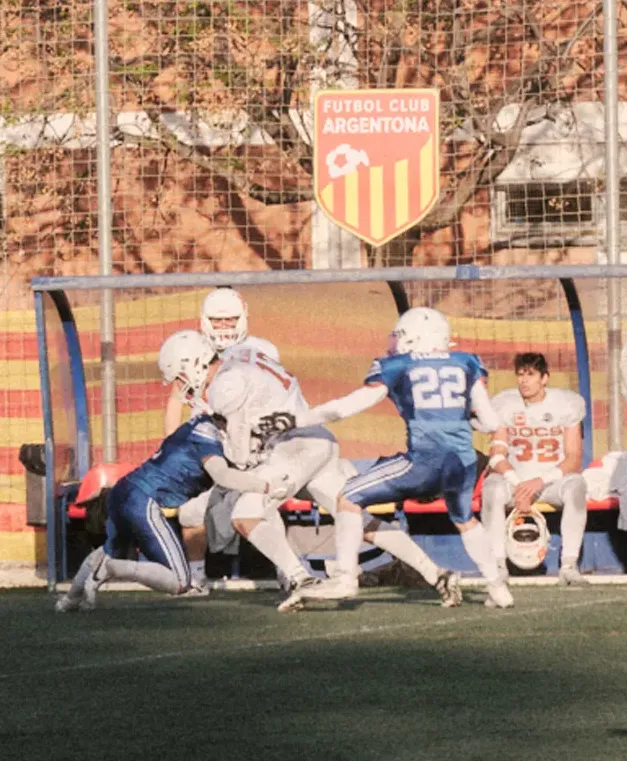 Close-up shot of a tackle near the sideline, showing players fighting for the ball in front of the team bench.