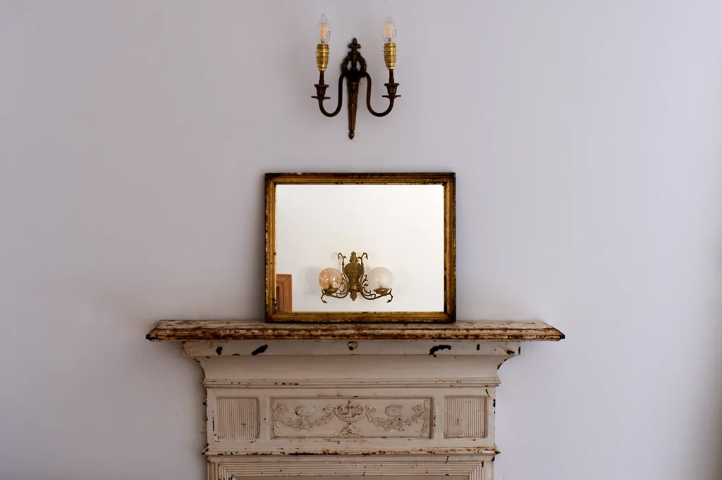 Ornate white fireplace mantel with distressed paint, topped with a rectangular antique mirror reflecting the room and an overhead brass wall sconce.
