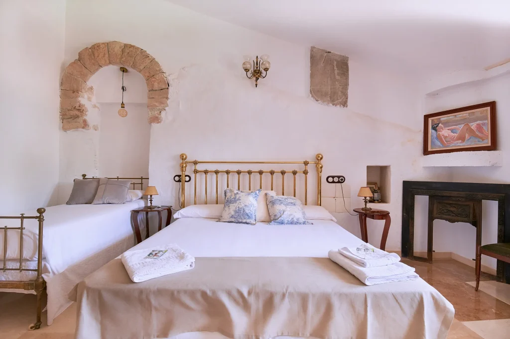Close-up of the master bed with a brass headboard, white linens, antique fireplace mantel, and a stone archway leading to a niche with a bed.