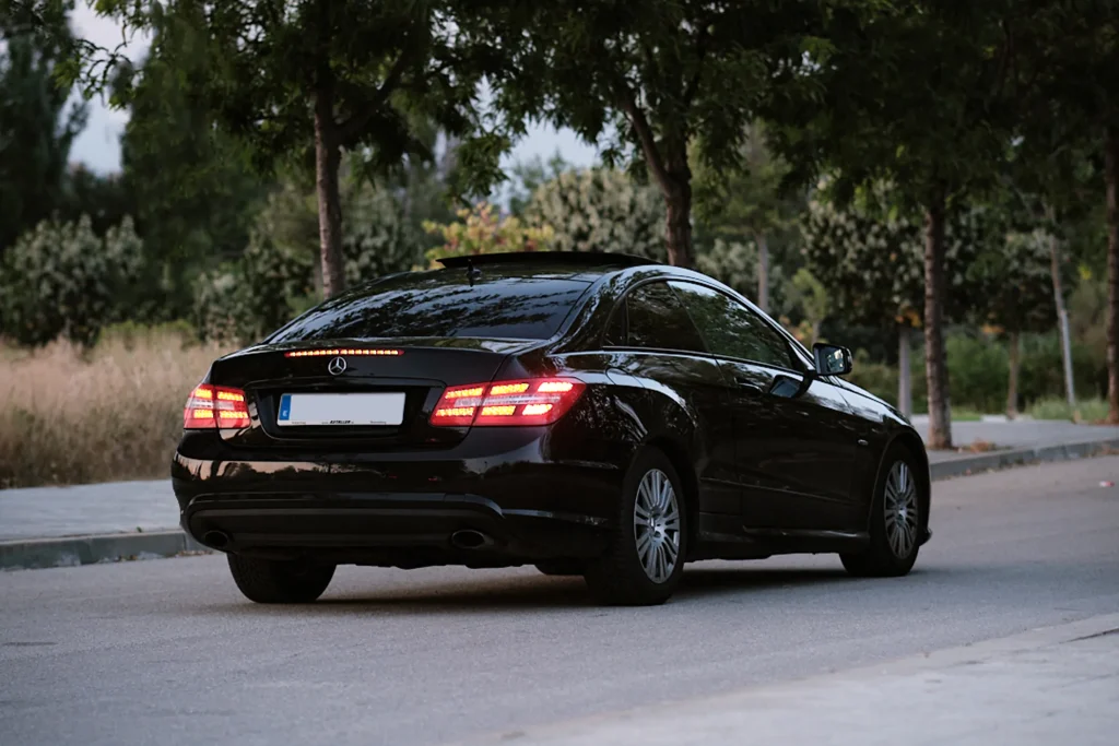 Dynamic rear three-quarter shot of the black Mercedes-Benz E-Class Coupé (C207) with illuminated LED taillights on a tree-lined street.