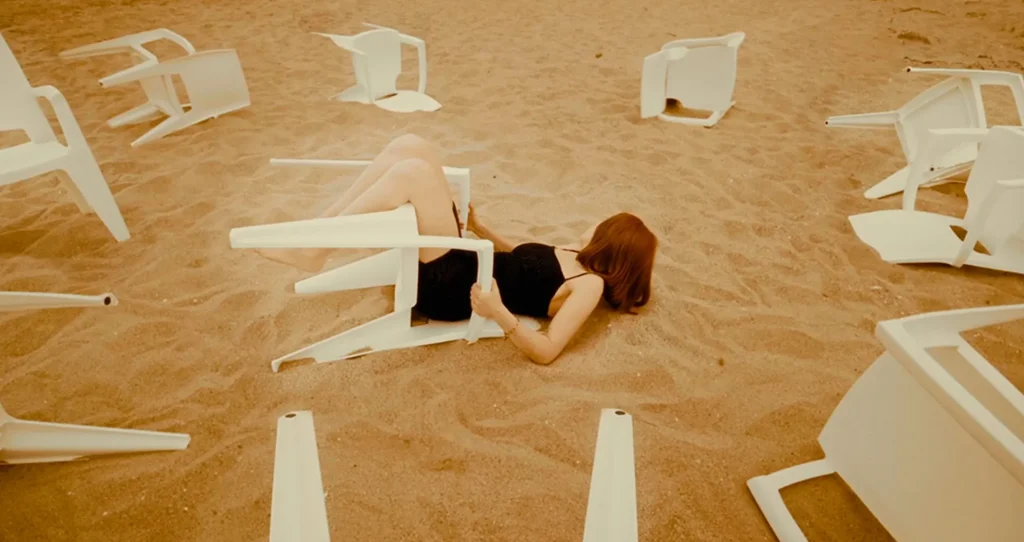 Surreal scene from the Burnout short film: woman lying amidst overturned white chairs on a sandy beach