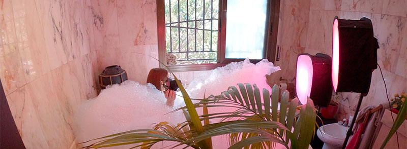 Making-of shot showing the actress in the bathtub surrounded by foam, with pink professional LED lights set up for the shoot.