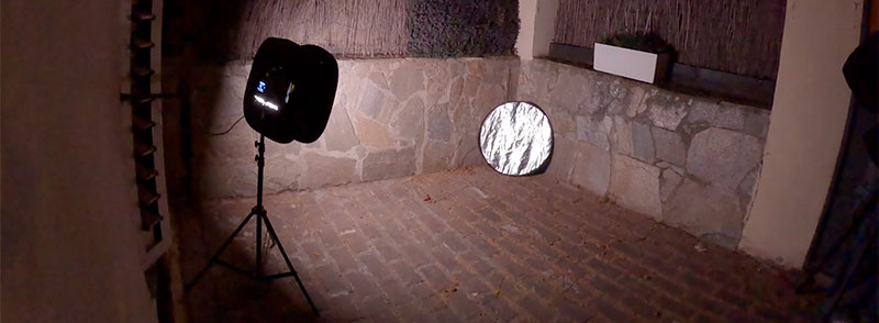 "Outdoor film production setup with a lighting stand and a silver reflector against a stone wall