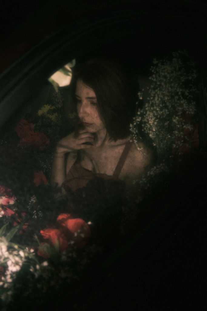 A pensive shot of the protagonist seen through the car window, surrounded by flowers, emphasizing a dreamy mood.