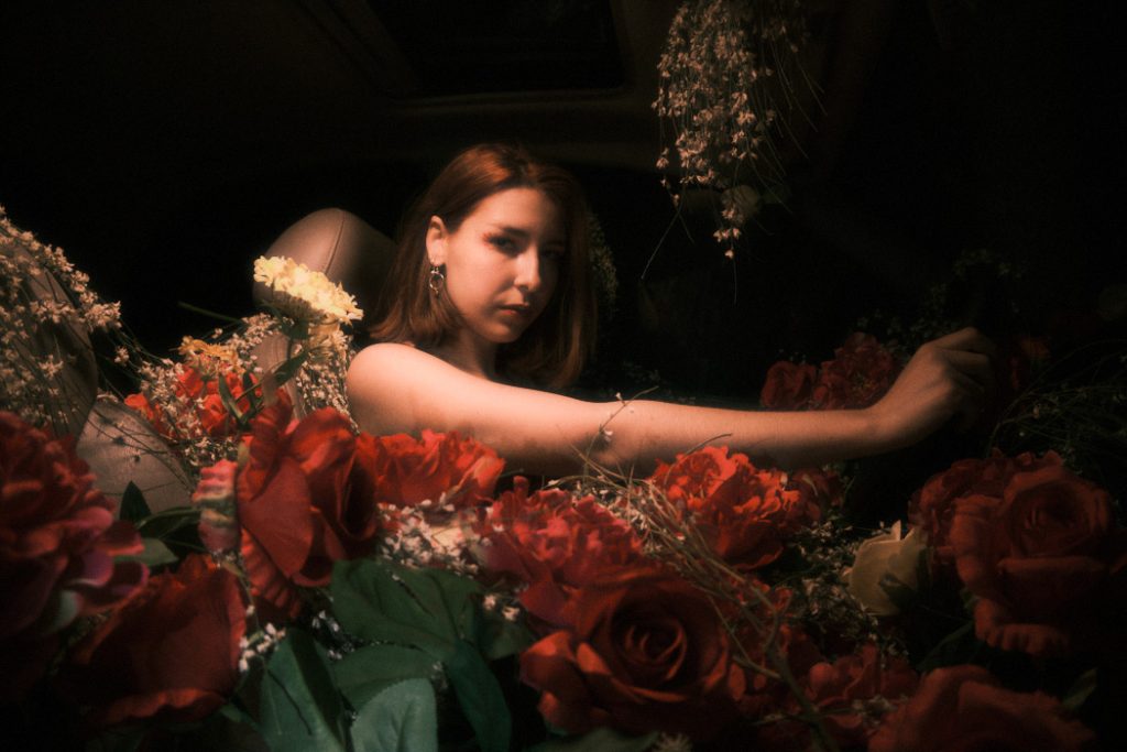 A moody portrait of the protagonist surrounded by deep red roses inside a dark car, stretching her arm.