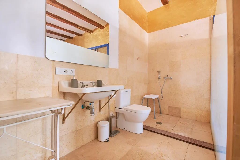 Tiled bathroom in Castell Granadella with a sink, toilet, walk-in shower, and a mirror reflecting the rustic wooden ceiling beams.