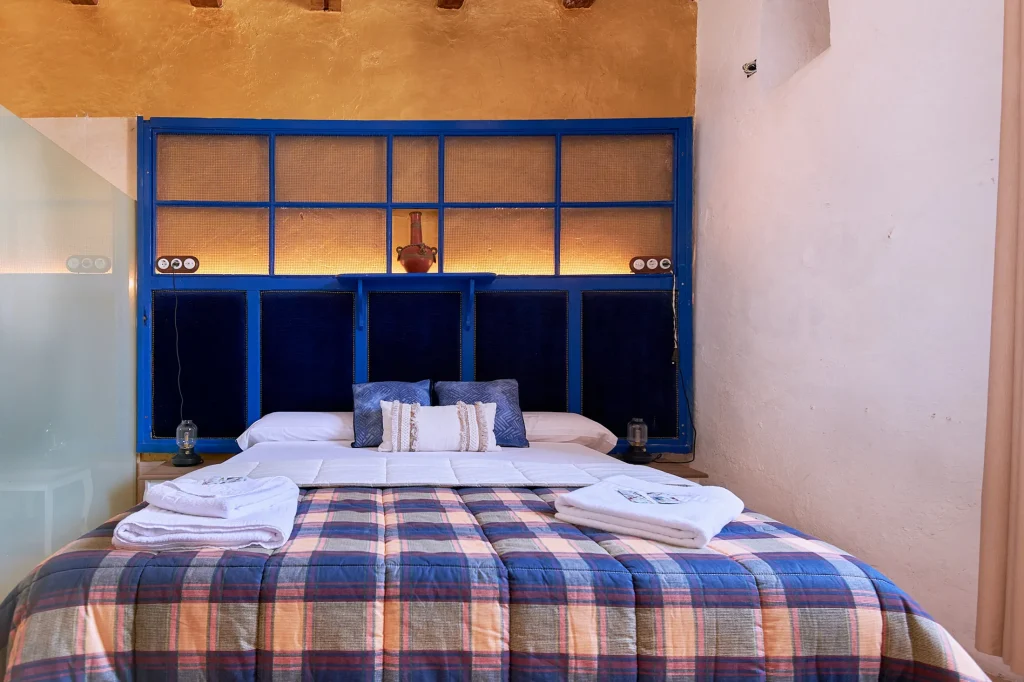Rustic interior design of a Castell Granadella apartment bedroom. Features a blue headboard with accent lighting, plaid bedding, and a full view of the main bed area.