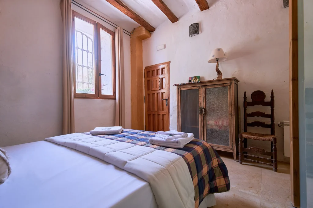 Second view of a bedroom in Castell Granadella. Shows a wooden vintage wardrobe with mesh panels, an antique chair, and a gridded window with neutral curtains.