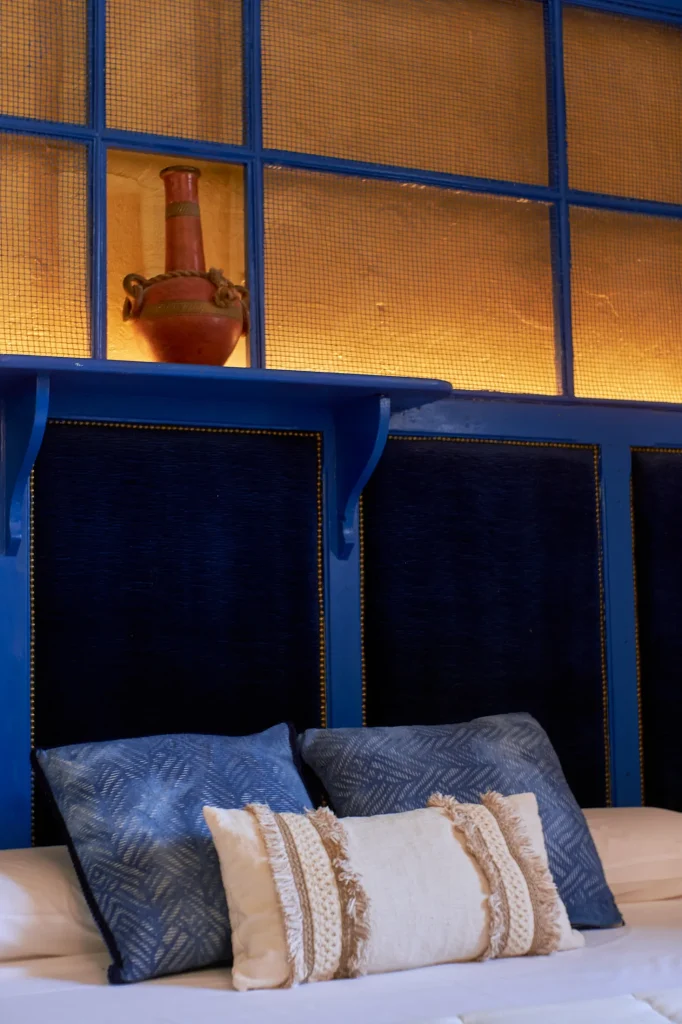 Rustic interior design of a Castell Granadella apartment bedroom. Features a blue headboard with accent lighting, plaid bedding, and a full view of the main bed area.