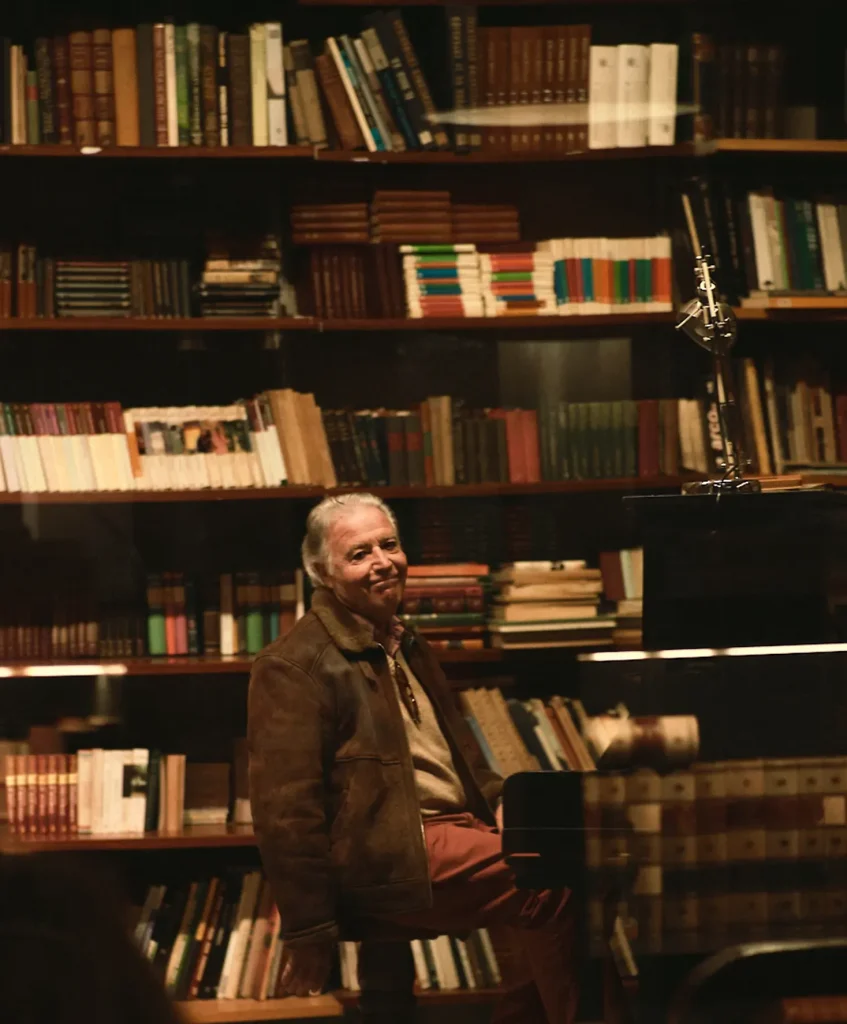 Cinematic and moody editorial portrait of an elder man in a brown jacket sitting in a library by a piano, shot with soft lighting for a high-end lifestyle feature or magazine content