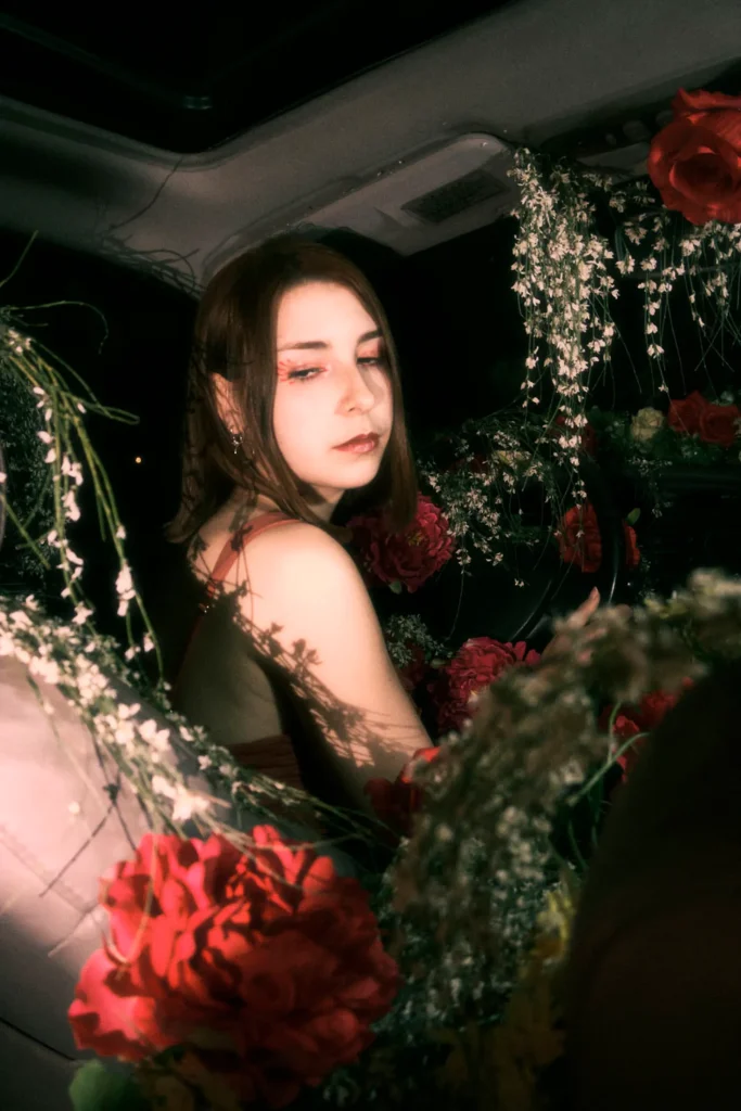 Conceptual fashion portrait of a woman surrounded by red and white roses in a car, with stylized makeup and low-key lighting for a surreal effect.