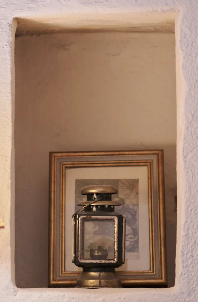 Detail shot of a white wall niche with an exposed texture, holding a framed picture and a small vintage lantern, beside a bedside lamp.