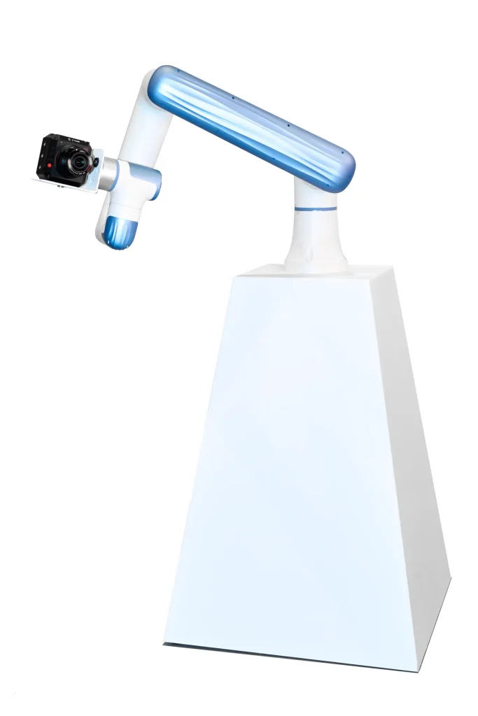 Close-up shot of a white and blue robotic arm (Glambot) holding a professional Canon camera, on a white pedestal.