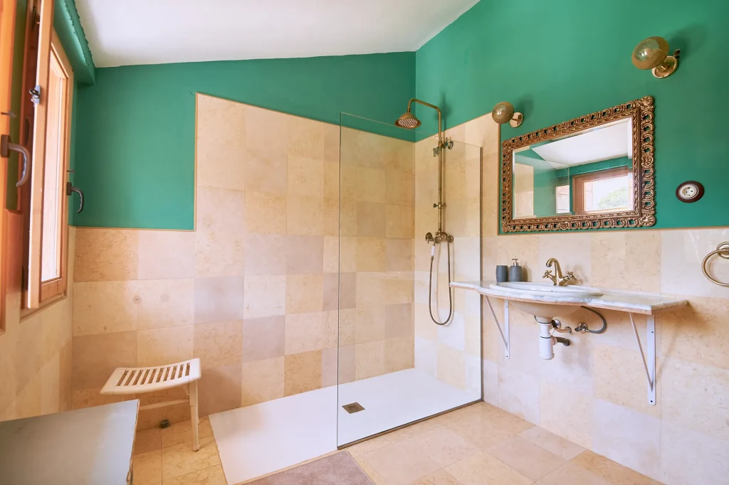 Designer bathroom with emerald green walls, beige tiling, a modern walk-in shower with brass fittings, and a decorative gold-framed mirror.