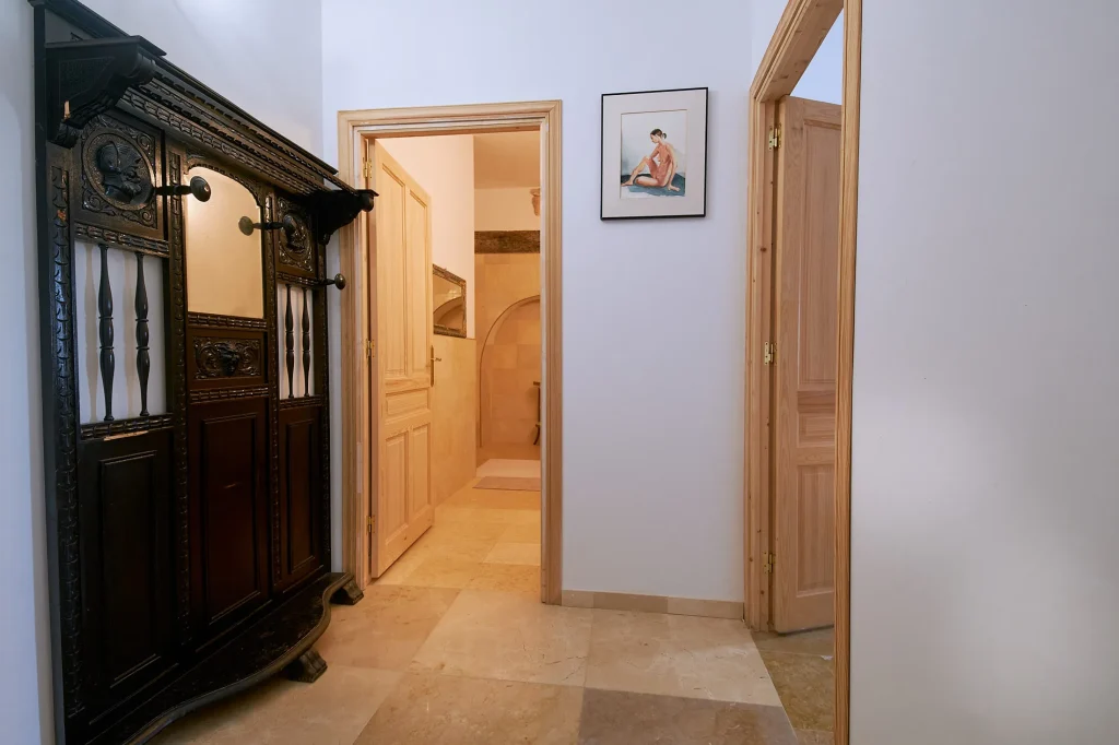 Entrance hallway with a dark, antique carved wooden coat rack (perchero), marble-tiled floor, and multiple light wooden doors leading to different rooms, including the bathroom.