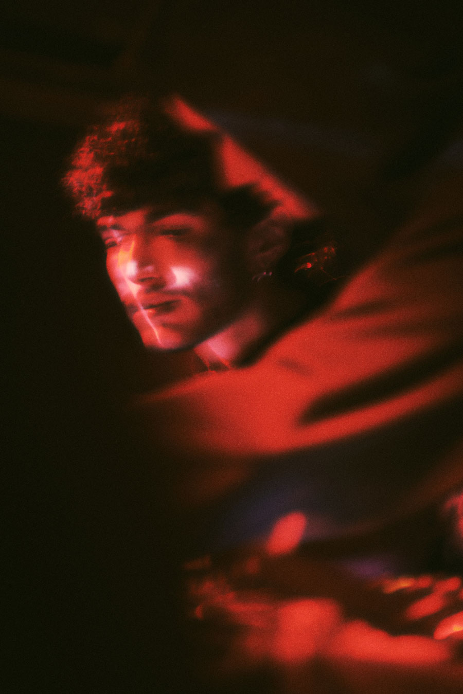 Documentary low-light photography from the RAIT music event, capturing a DJ/artist portrait with dramatic red lighting and cinematic grain for European event promotion.