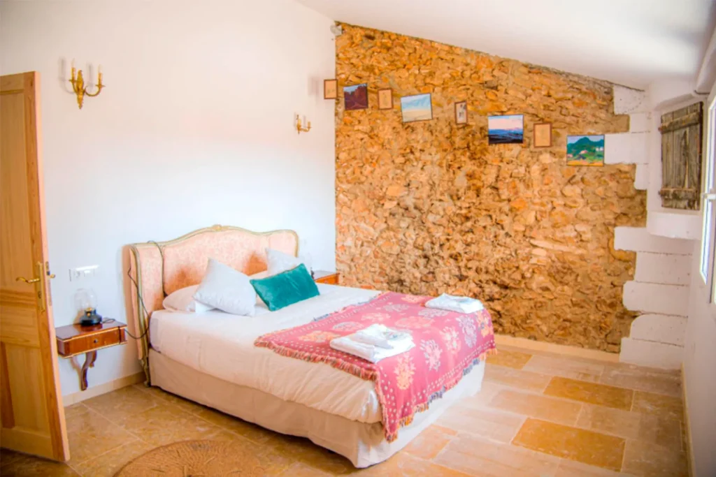 Previous photo (low quality) of a rustic bedroom with an exposed stone wall, a pink/beige upholstered bed, and a colorful throw blanket. IMPORTANTE: Usa esto para las galerías del "Antes"