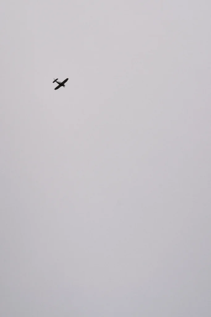 Minimalist, high-key conceptual travel photograph of a small airplane silhouette against a vast, bright grey sky, representing the journey or travel destination